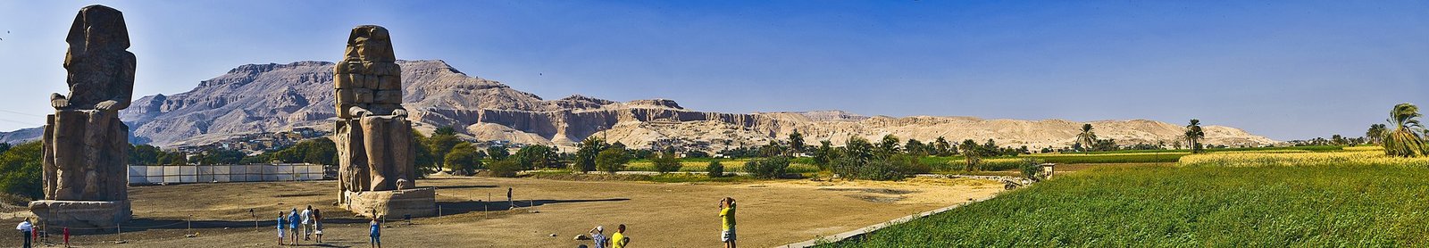 A panoramic view of the Colossi of Memnon, two colossal statues of Pharaoh Amenhotep III, standing in the Valley of the Kings, Luxor, Egypt. The statues are surrounded by the desert landscape and the Nile River is visible in the background.