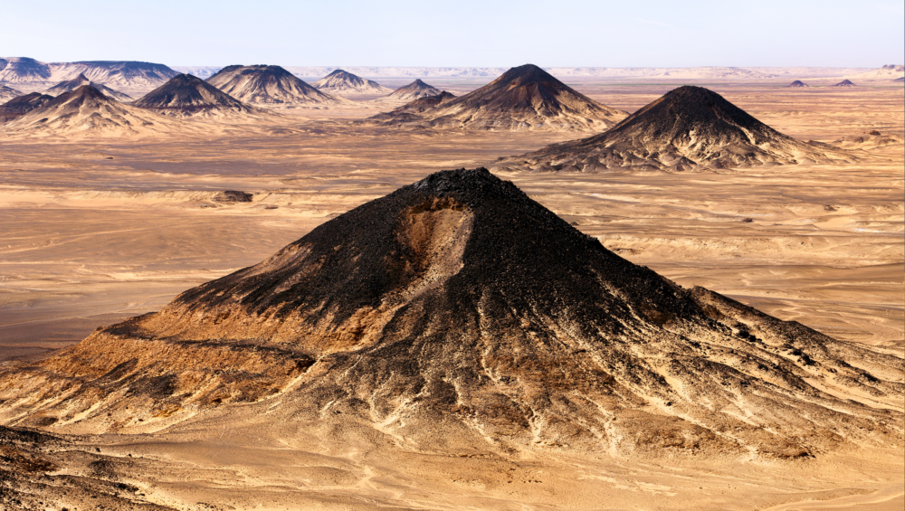 A panoramic view of the Black Desert in Egypt, showcasing the black volcanic mountains, the sand dunes, and the clear blue sky.