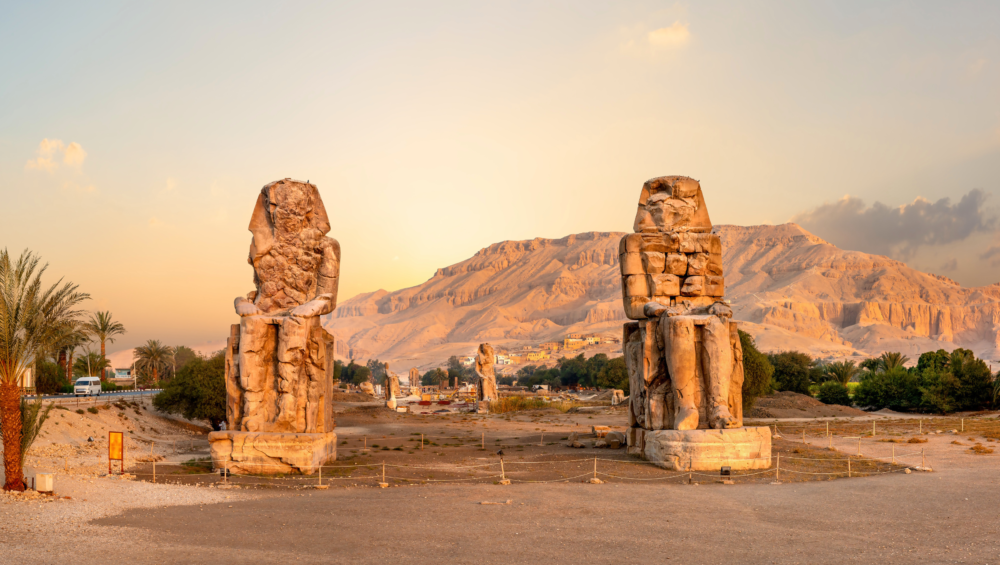 A photo of the Colossi of Memnon, two colossal statues of Pharaoh Amenhotep III, standing in the Valley of the Kings, Luxor, Egypt. The statues are surrounded by the desert landscape and the Nile River is visible in the background.