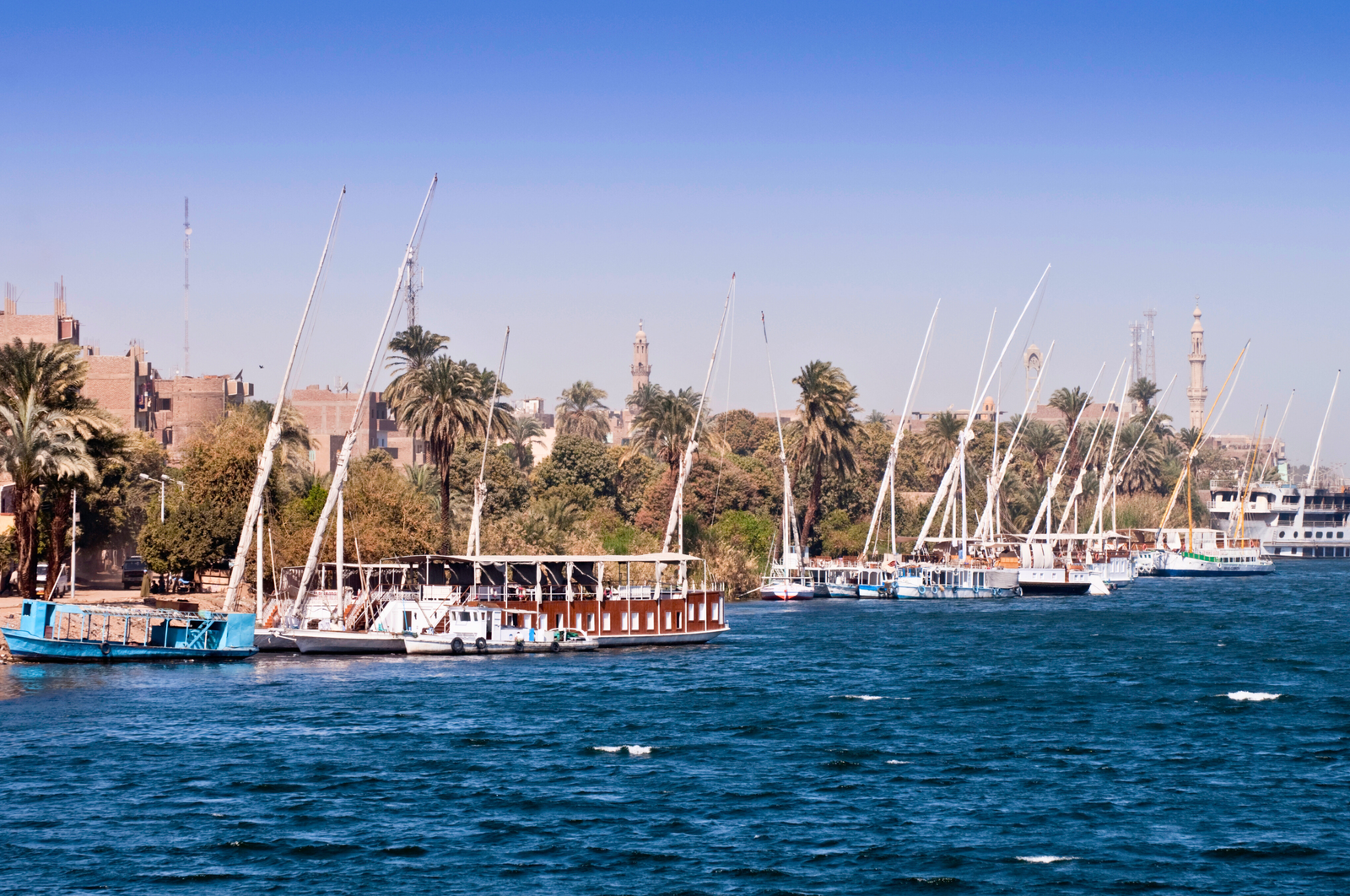 A panoramic view of the Nile River in Luxor, Egypt, featuring feluccas and a dahabiya sailing on the water, palm trees lining the riverbank, and ancient structures in the background.