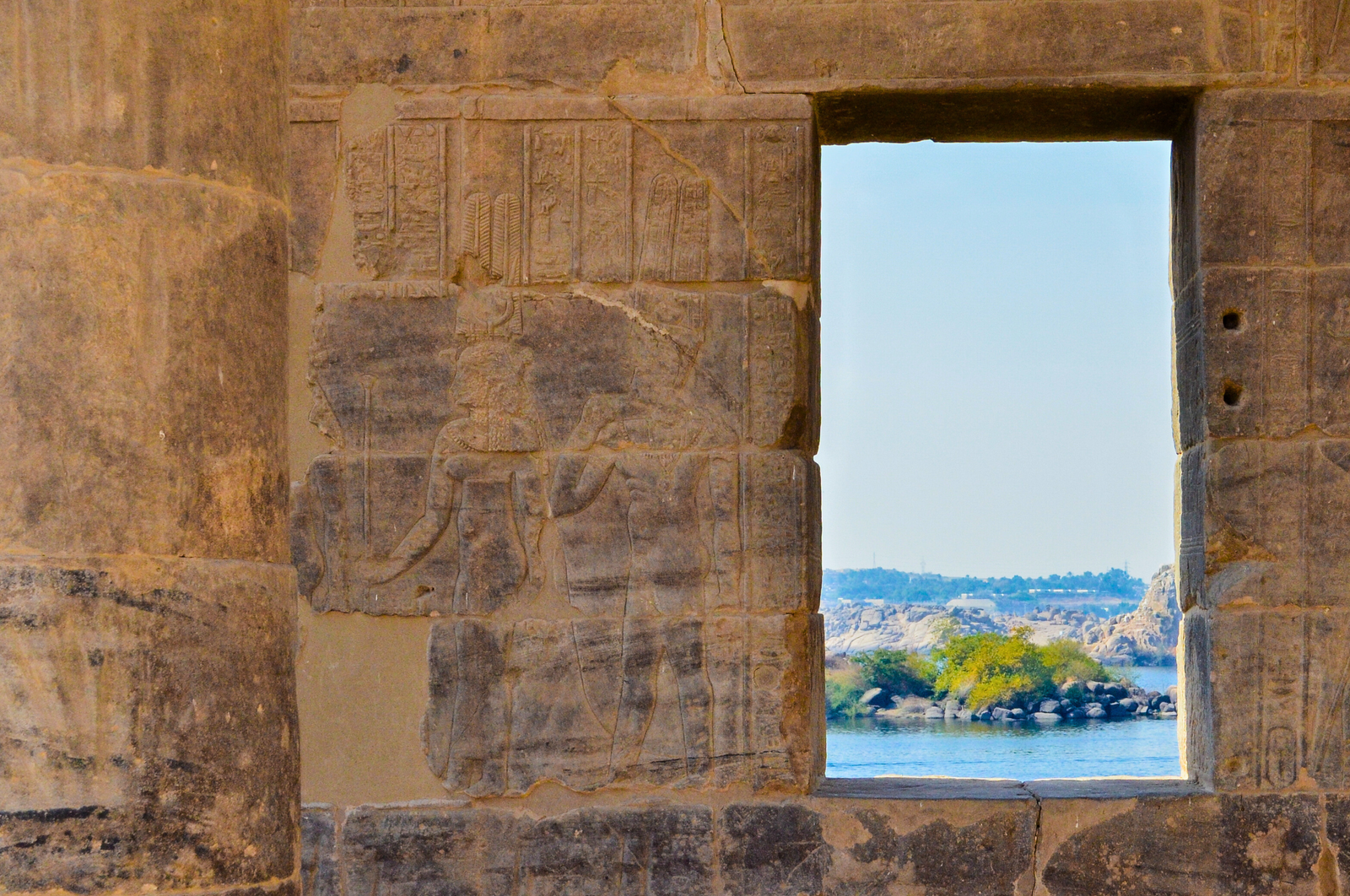 A photo of a window in Philae Temple, showcasing a view of the Nile River with a small island in the background. The window frame is decorated with intricate hieroglyphs and carvings.