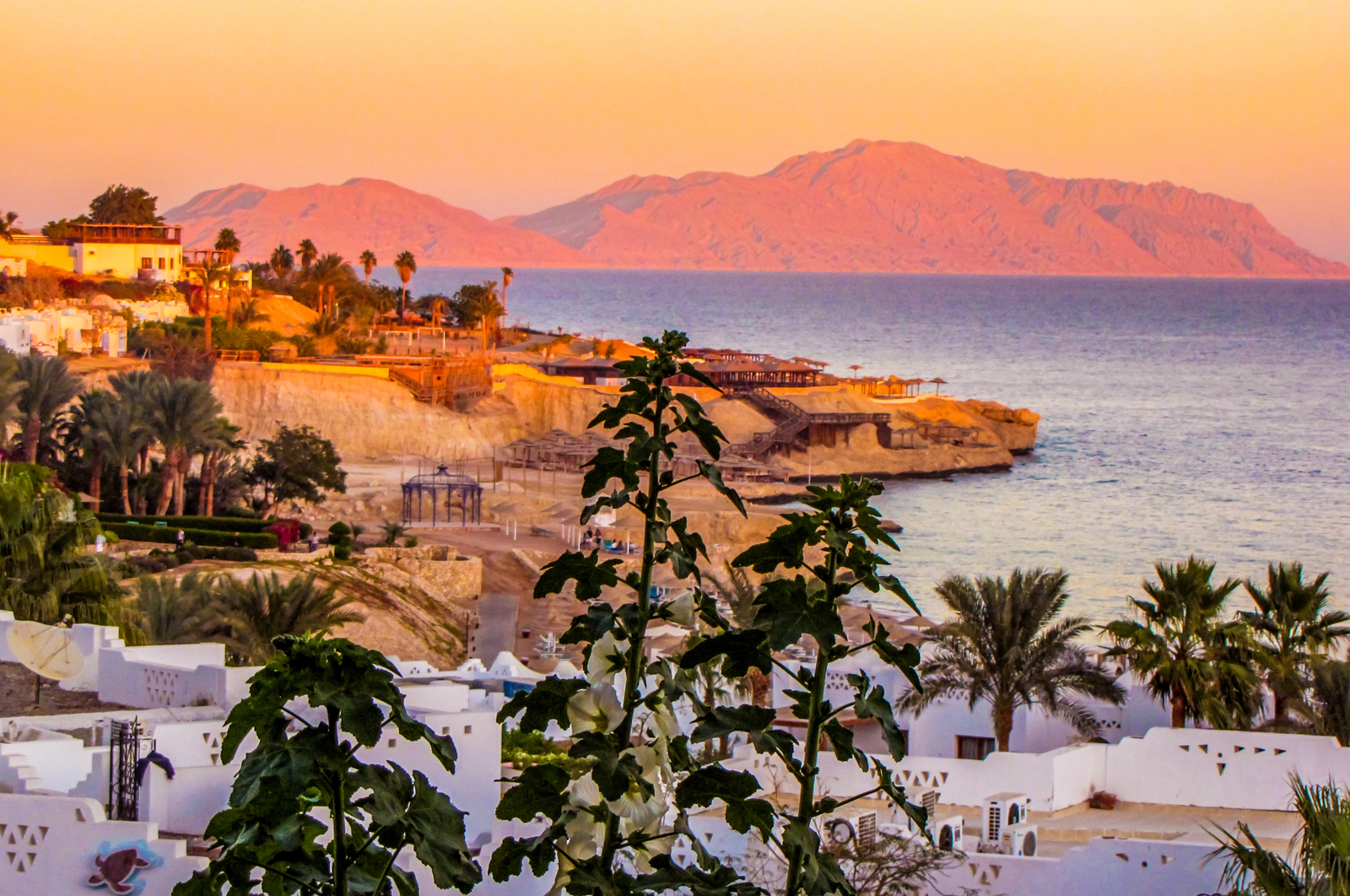 A panoramic view of Sharm El Sheikh, Egypt, at sunset, showcasing the red mountains, the coastal town with white buildings and palm trees, and the calm Red Sea.