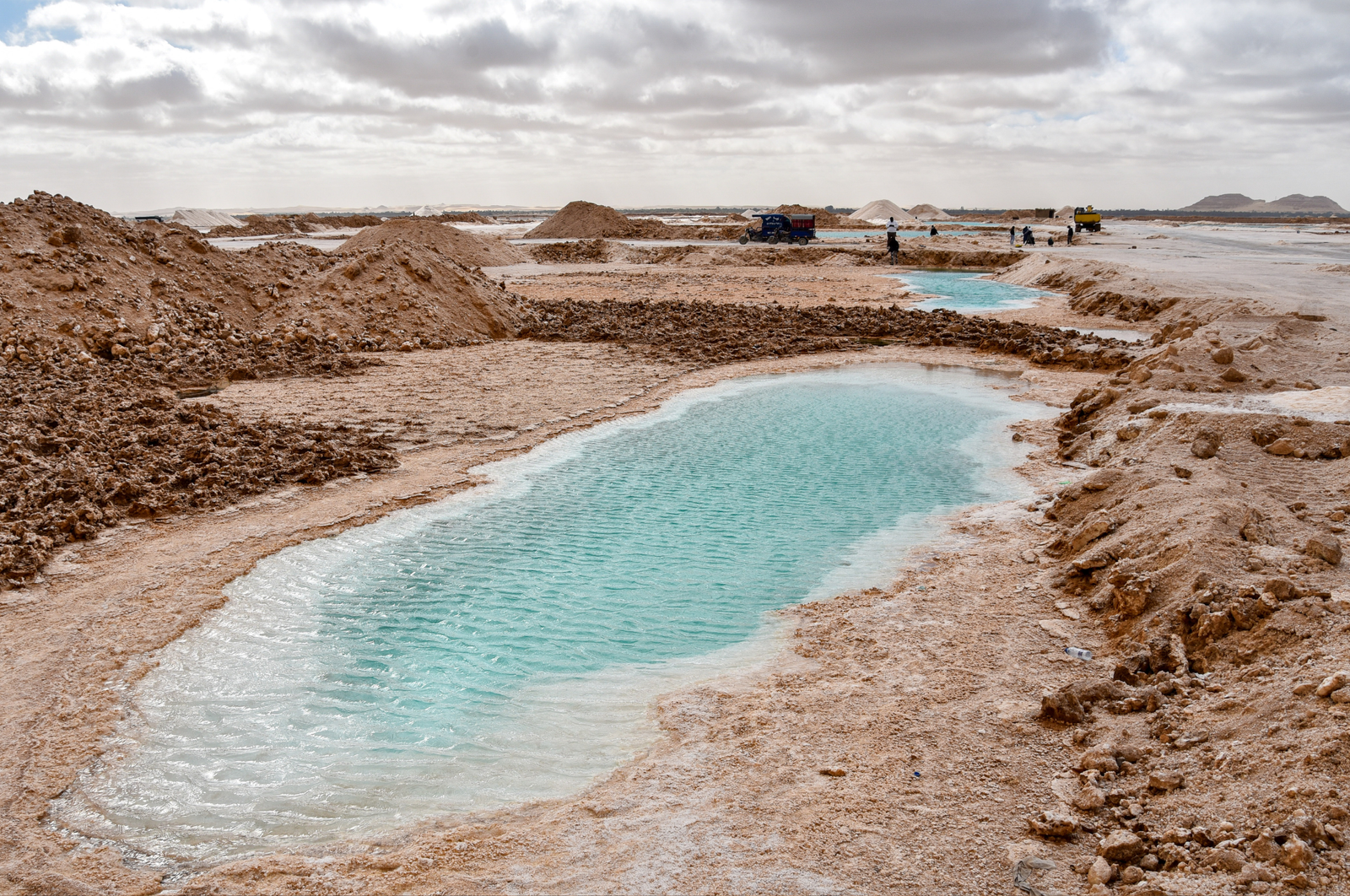  A panoramic view of the Siwa Oasis in Egypt, showcasing salt pans, mining operations, and a small pool of turquoise water.