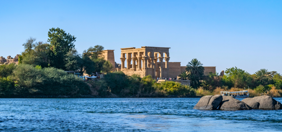 Philae Temple viewed from the Nile, surrounded by water and lush vegetation.