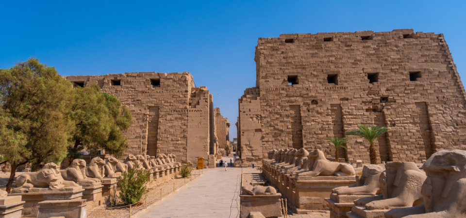 A photo of the Avenue of Sphinxes at Karnak Temple, showcasing rows of ram-headed sphinxes leading towards the towering temple pylons.