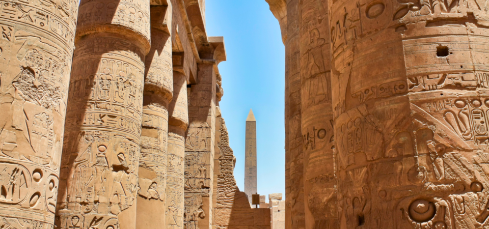 A photo of the towering columns in Karnak Temple's Hypostyle Hall, with a distant obelisk visible.