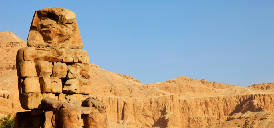 A photo of the Colossi of Memnon, two colossal statues of Pharaoh Amenhotep III, standing in the Valley of the Kings, Luxor, Egypt. The statues are surrounded by the desert landscape and the Nile River is visible in the background.