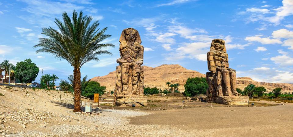 Two colossal statues of Pharaoh Amenhotep III, known as the Colossi of Memnon, standing in the Valley of the Kings, Luxor, Egypt. A palm tree stands in the foreground.