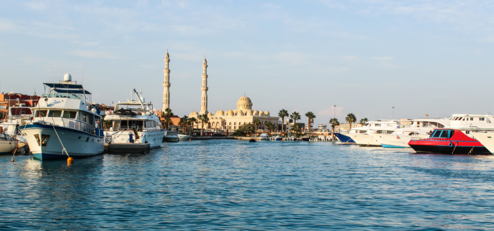 A panoramic view of the Hurghada Marina, featuring yachts docked in the harbor, palm trees lining the waterfront, and Al Mina Mosque in the background.