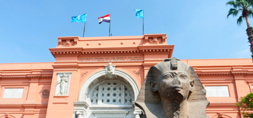 A photo of the Egyptian Museum in Cairo, featuring a large statue of a sphinx in front of the building, with Egyptian flag flying above.