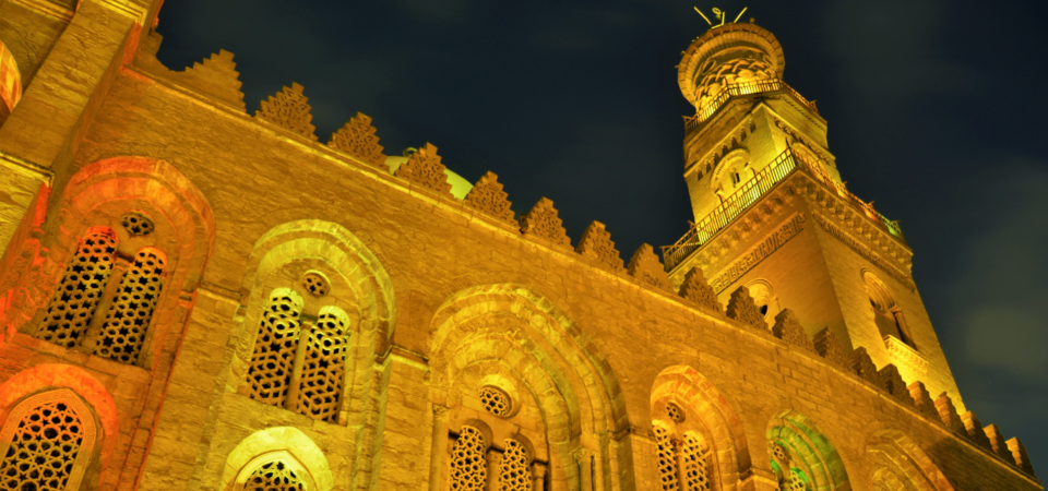 A close-up night view of the Qalawun Complex in Cairo, Egypt, highlighting its illuminated facade, intricate carvings, and towering minaret.