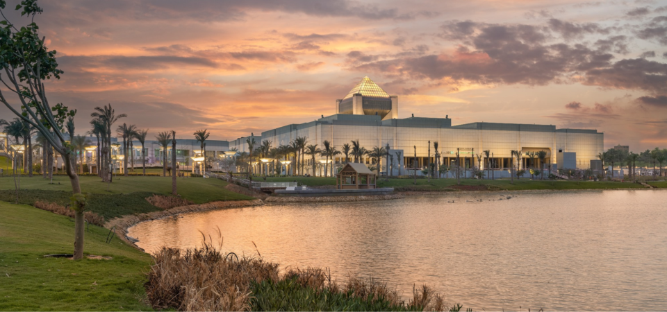 A modern museum building with a pyramid-shaped roof, surrounded by a lake and palm trees, against a dramatic sunset sky.