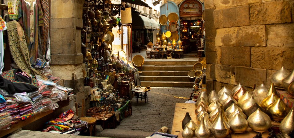 A narrow alleyway in Khan el-Khalili, Cairo, filled with shops selling colorful textiles, handicrafts, and metalwork.