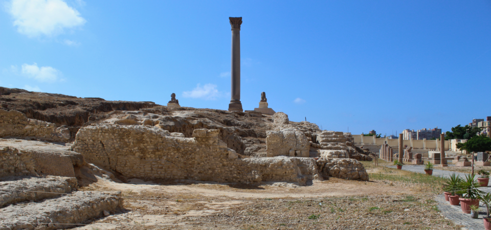 A photo of Pompey's Pillar in Alexandria, Egypt, a tall granite column standing amidst ancient ruins.