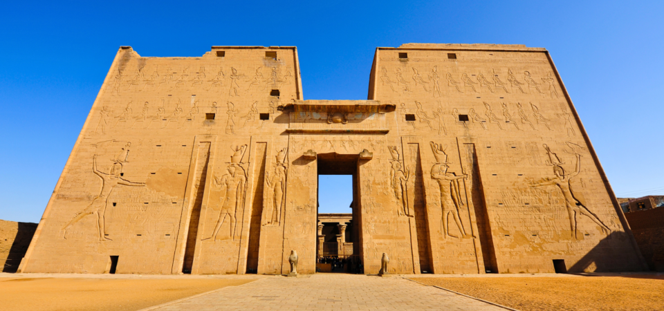 A photo of the entrance to the Temple of Horus in Edfu, Egypt, showcasing the massive pylons, decorated with hieroglyphs, and the columns leading into the temple.