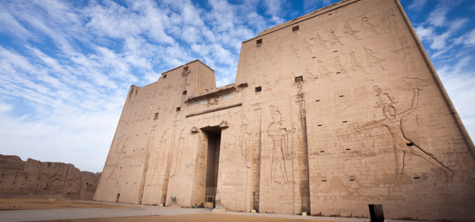 A massive, towering pylon entrance to the Temple of Horus in Edfu, Egypt, with intricate hieroglyphs and reliefs.