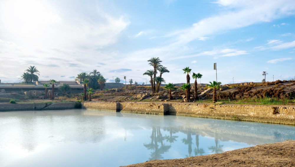 A photo of the Sacred Lake at Karnak Temple, showing the calm water, palm trees, and ancient temple ruins.