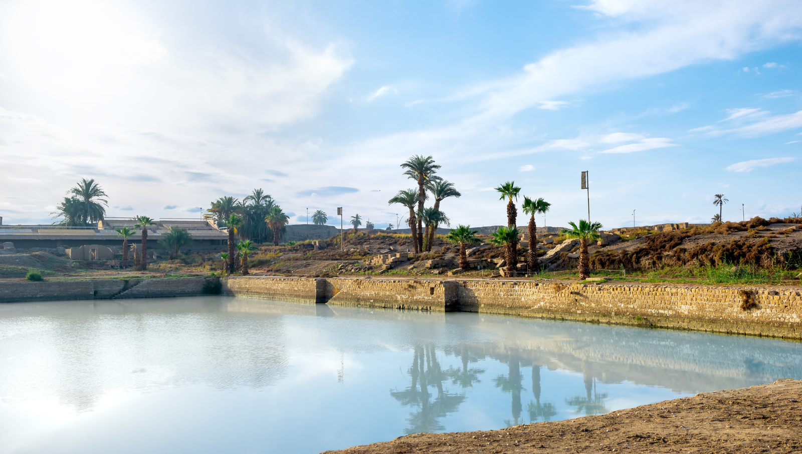 A photo of the Sacred Lake at Karnak Temple, showing the calm water, palm trees, and ancient temple ruins.