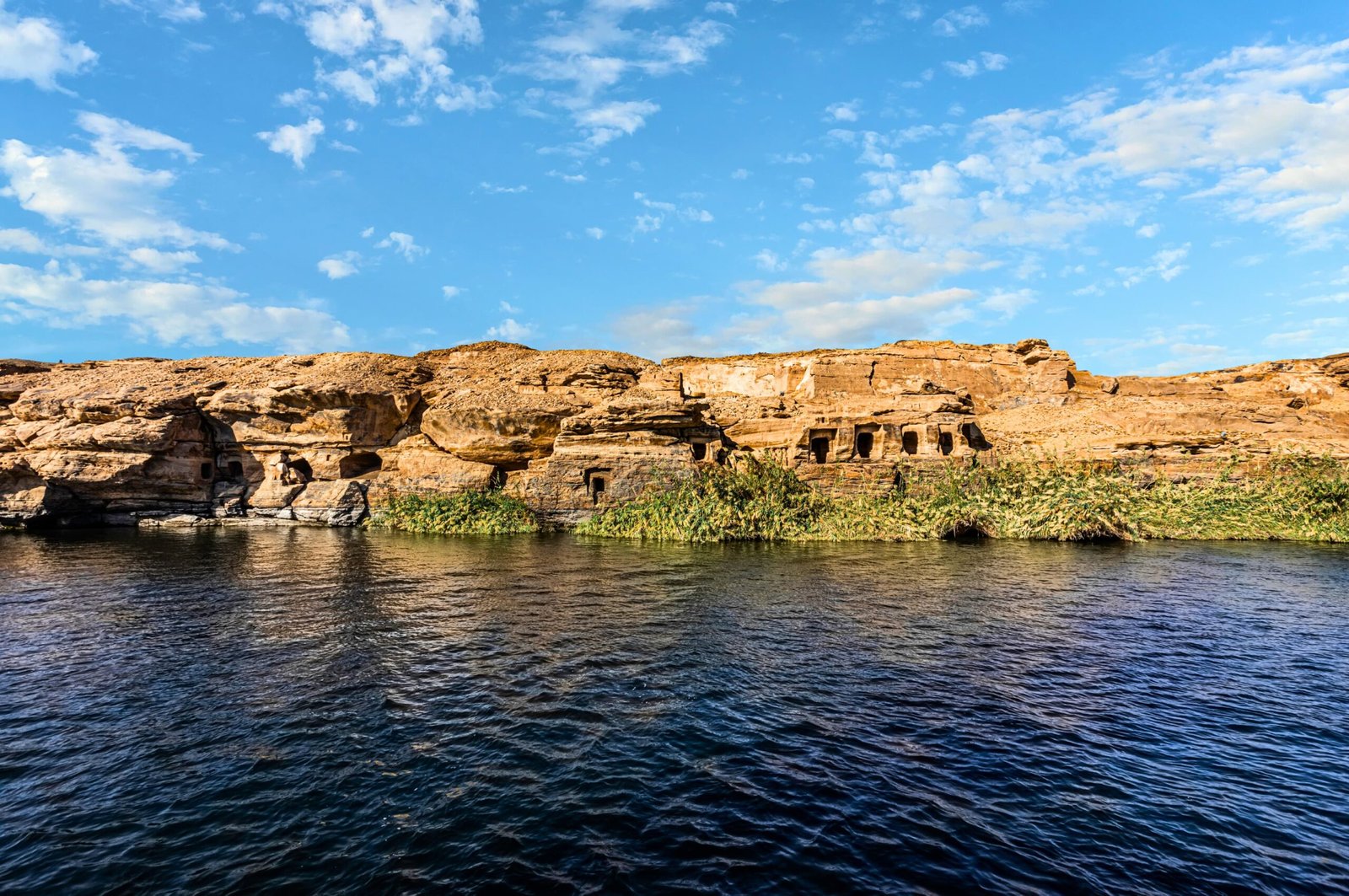 A photo of Gebel el-Silsila, showcasing the sandstone cliffs carved with ancient tombs and the Nile River flowing in the foreground.