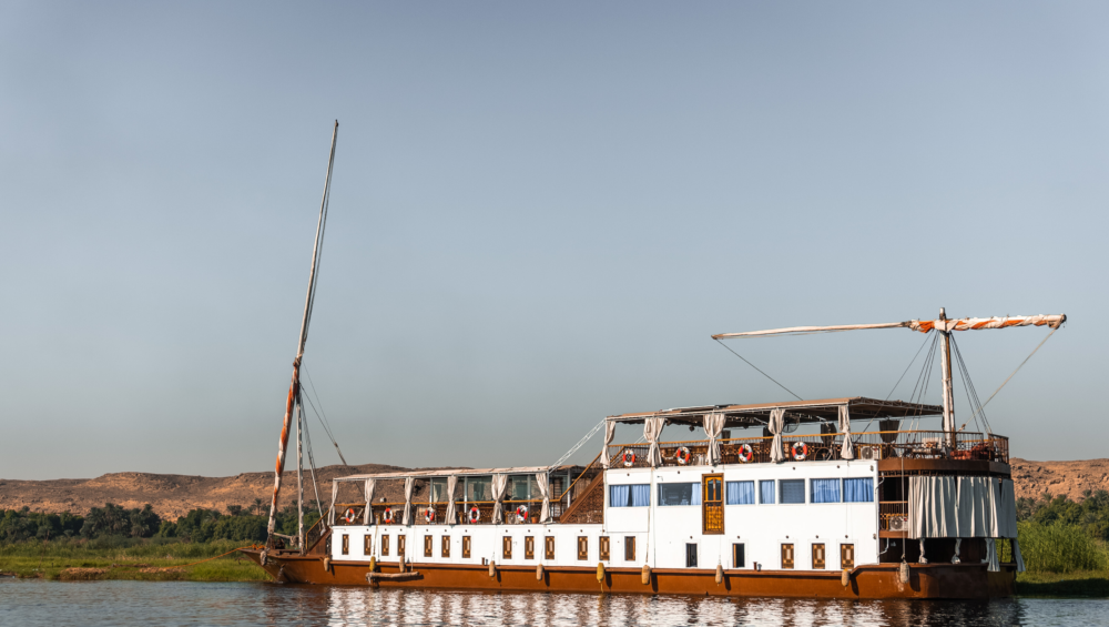 A traditional Egyptian dahabiya sailing boat on the Nile River, with white sails and a wooden hull.