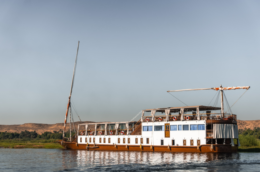 A traditional Egyptian dahabiya sailing boat on the Nile River, with white sails and a wooden hull.