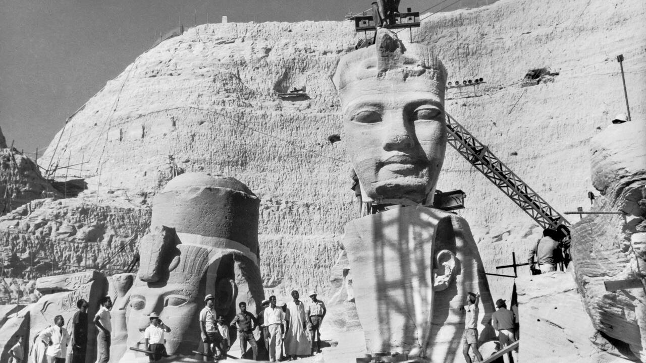  A black and white photo of the Great Temple of Abu Simbel during its relocation project, showing the colossal statues being dismantled and lifted by cranes.