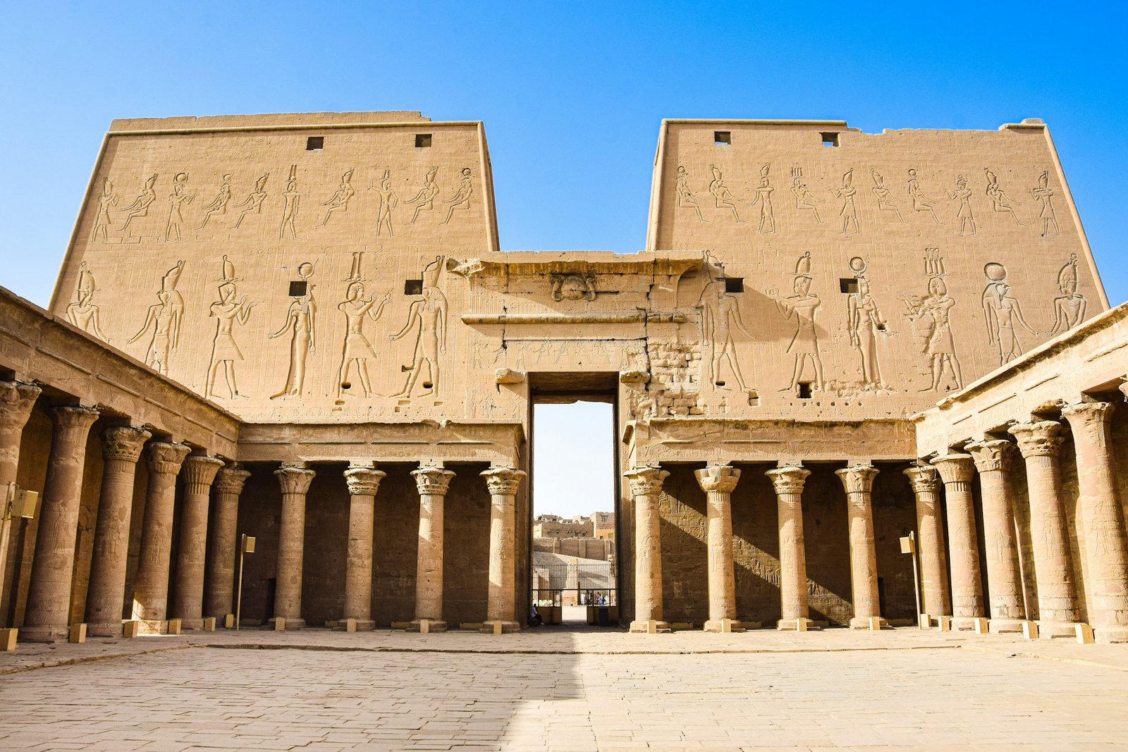 A photo of the facade of the Temple of Edfu, showcasing the massive pylons, decorated with hieroglyphs, and the columns leading into the temple.