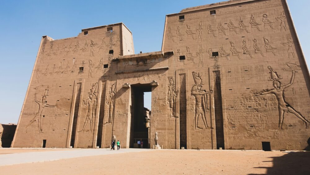 A photo of the facade of the Temple of Edfu, showcasing the massive pylons, decorated with hieroglyphs, and the columns leading into the temple.