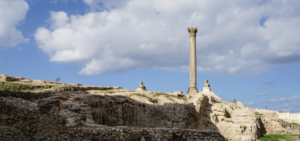 Pompey’s Pillar in Alexandria standing tall amidst ruins, flanked by two sphinx statues under a partly cloudy sky.