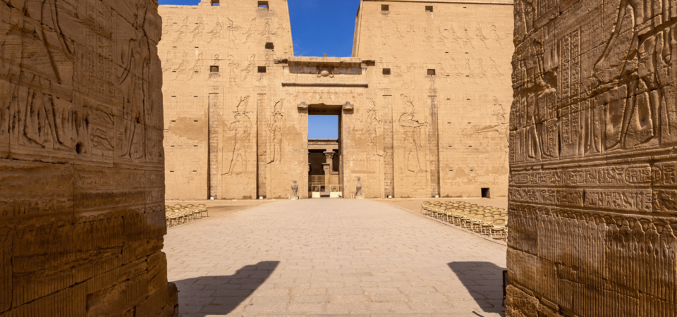 The grand entrance of Temple of Horus (Edfu Temple), flanked by towering pylons adorned with carvings, under a vivid blue sky.