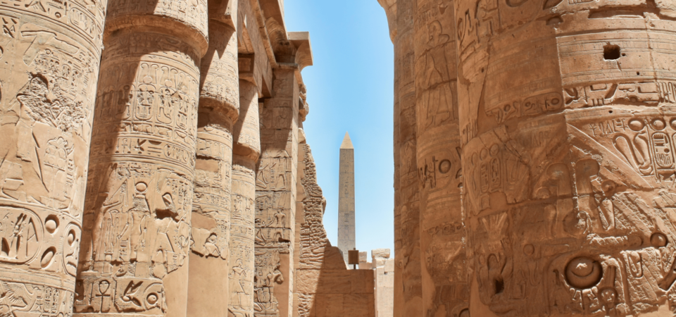 Massive columns of Karnak Temple with intricate hieroglyphs and an obelisk in the background.