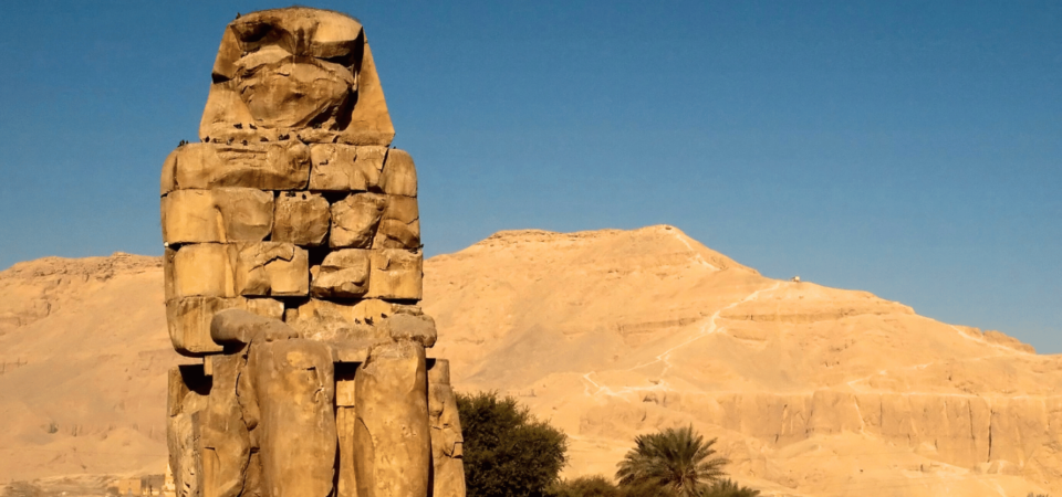 One of the colossal statues of the Colossi of Memnon against a backdrop of rugged mountains and a clear sky.