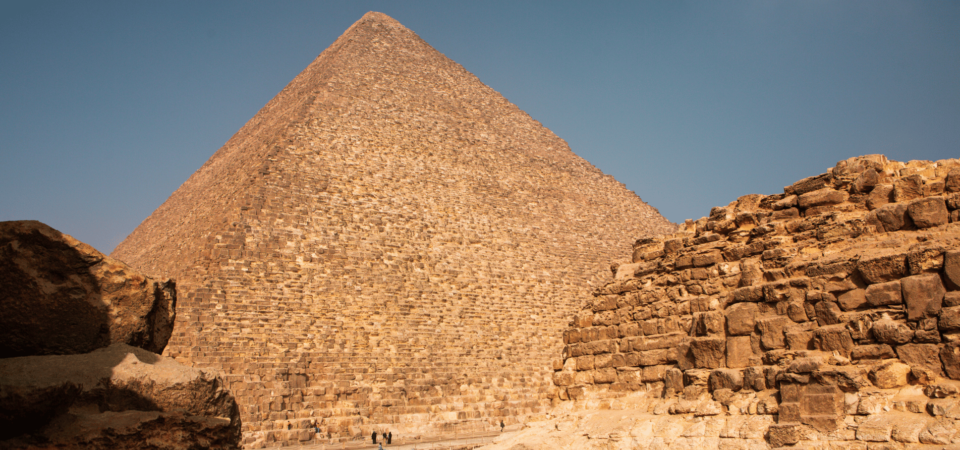 The Great Pyramid of Giza towering under the blue sky, with surrounding ancient stones in the foreground.